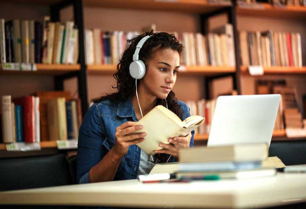 woman with headphones sitting at library table looking at laptop while holding an open book